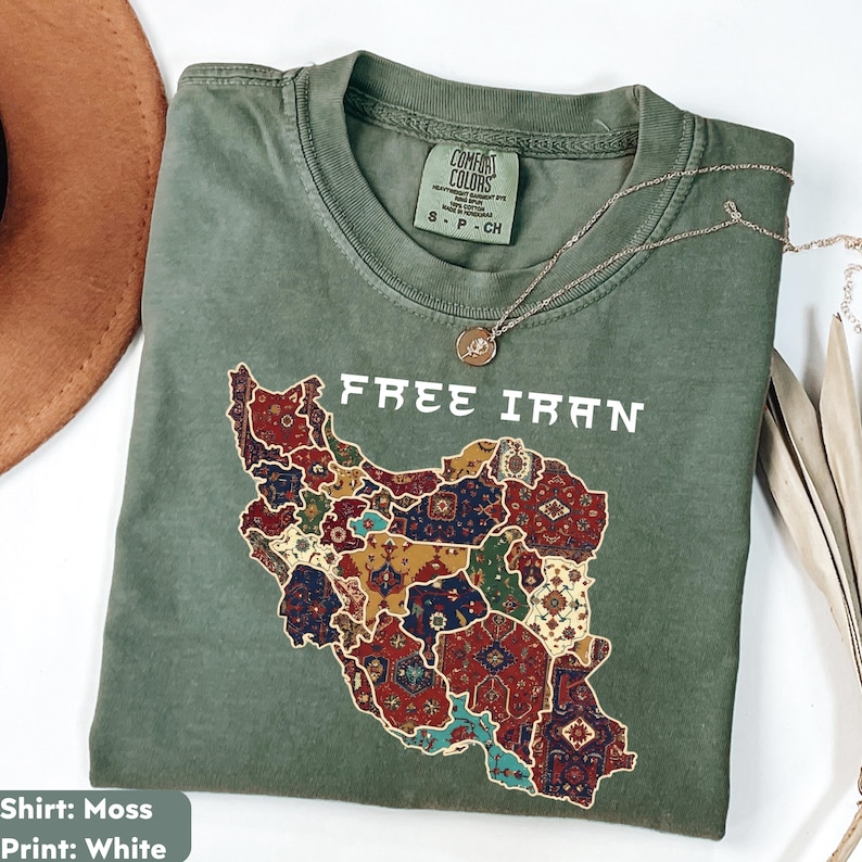 以下が含まれることがあります： モスグリーンのTシャツ。前面には「FREE IRAN」の文字と、カラフルな模様で埋められたイランの地図がプリントされています。シルバーのネックレスがTシャツの上に置かれています。Tシャツには「Comfort Colors」と記載されています。