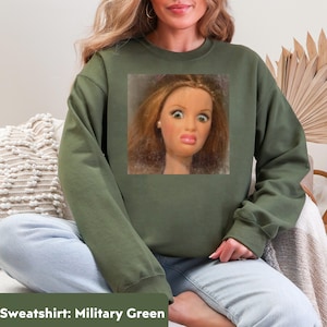 May include: Military green sweatshirt featuring a doll's face with a surprised expression. The doll has red hair, large eyes, and a pink mouth. The sweatshirt is a solid color and has a crew neck.