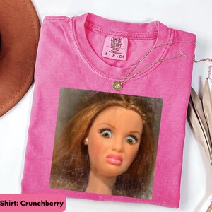 May include: A pink Comfort Colors t-shirt with a doll face graphic. The shirt has a crew neck and short sleeves. A gold necklace with a pendant is draped over the shirt. The shirt is labeled "Crunchberry".