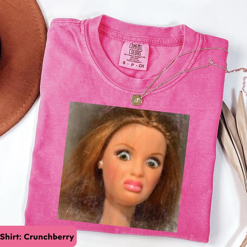 May include: A pink Comfort Colors t-shirt with a doll face graphic. The shirt has a crew neck and short sleeves. A gold necklace with a pendant is draped over the shirt. The shirt is labeled "Crunchberry".