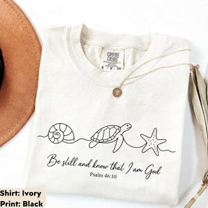 May include: An ivory t-shirt featuring a black line art graphic of a seashell, a sea turtle, and a starfish connected by a single continuous line. Below the graphic, the text reads, "Be still and know that I am God" and "Psalm 46:10".