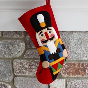 May include: Red felt Christmas stocking with a nutcracker design. The nutcracker is holding a drum and has a blue, yellow, and green uniform.