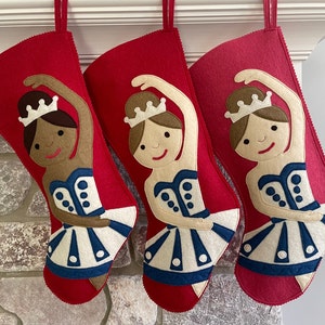 Könnte beinhalten: Drei rote Filz-Weihnachtsstrümpfe mit Ballerina-Motiven. Jeder Strumpf zeigt eine Ballerina mit einem weißen Tutu, einem blauen Oberteil und einer Krone. Die Ballerinas haben unterschiedliche Hauttöne und Haarfarben.