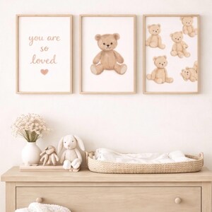 May include: Three framed prints with watercolor teddy bear illustrations and the text "you are so loved". A wooden dresser displays a woven changing basket, stuffed animals, and a vase of flowers. A white blanket is draped over the dresser.