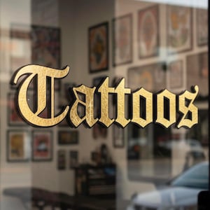 May include: A storefront window with the word "Tattoos" in gold lettering. The letters have a textured, gilded appearance and are set against a dark background. The reflection in the glass shows the interior of a tattoo parlor.