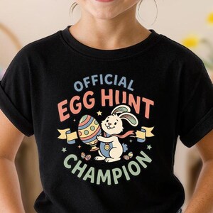 May include: A black t-shirt with the text "OFFICIAL EGG HUNT CHAMPION" and a graphic of a bunny holding an Easter egg. The shirt is worn by a child wearing sunglasses and a pink headband. Easter-themed decorations are in the background.