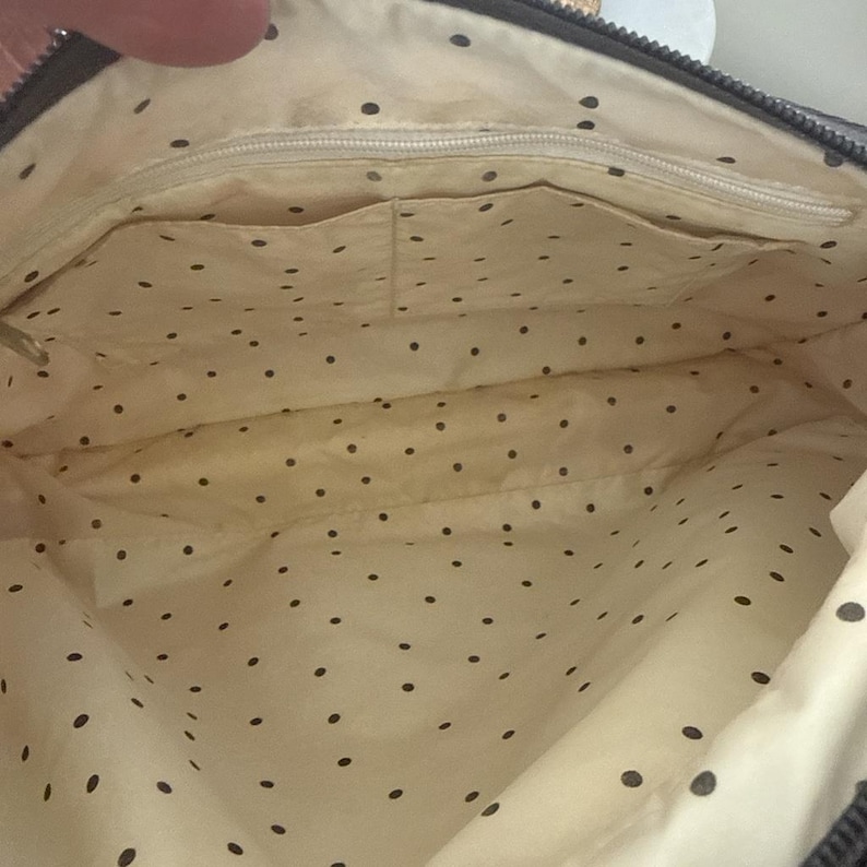 May include: Interior view of a dark gray handbag. The inside lining is cream-colored with small, evenly spaced black polka dots. The bag has several pockets and a zipper closure.