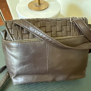 May include: A brown leather crossbody bag with a woven top flap. The bag has a rectangular shape with a long adjustable strap. The bag has a small pocket on the back and a flap closure. The bag is made of a soft, supple leather.