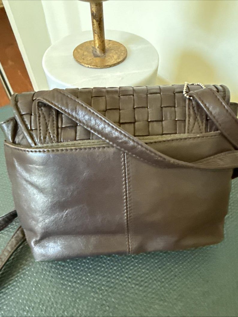 May include: A brown leather crossbody bag with a woven top flap. The bag has a rectangular shape with a long adjustable strap. The bag has a small pocket on the back and a flap closure. The bag is made of a soft, supple leather.