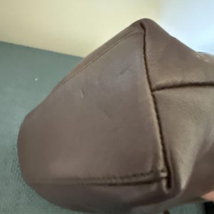 May include: A close-up of a brown leather handbag. The bag has a rounded bottom and a structured design. The stitching is visible along the seams, and the leather has a smooth texture. The bag appears to be new.