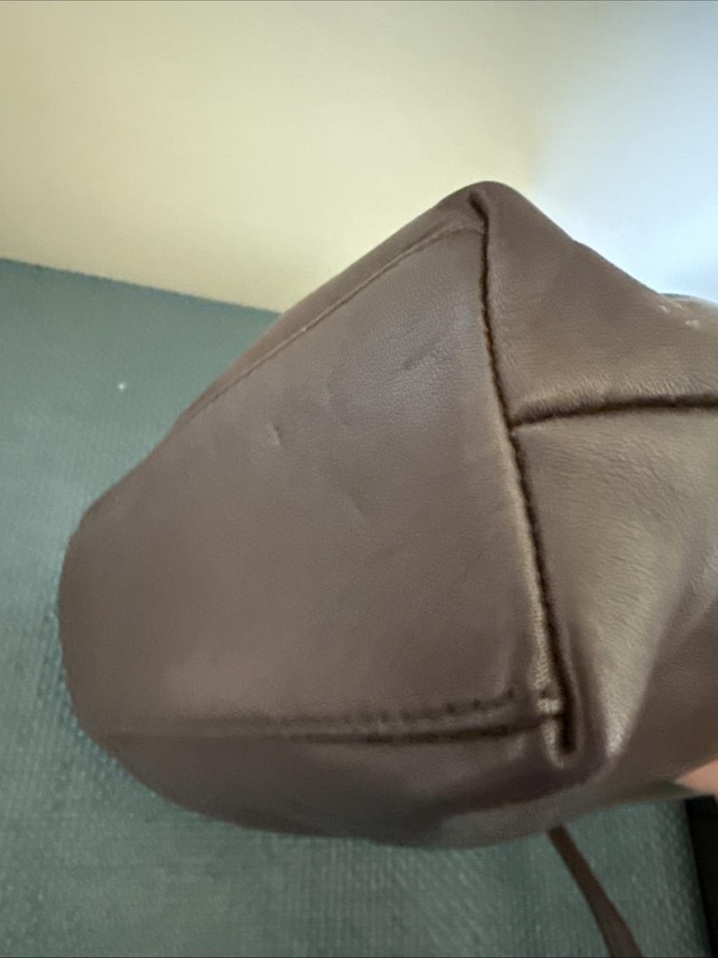 May include: A close-up of a brown leather handbag. The bag has a rounded bottom and a structured design. The stitching is visible along the seams, and the leather has a smooth texture. The bag appears to be new.