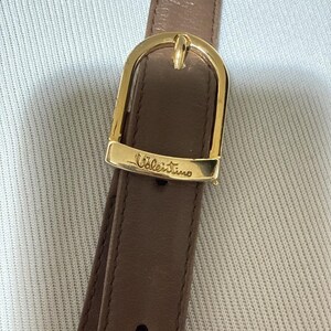 May include: A brown leather strap featuring a polished gold-tone metal buckle. The buckle is engraved with the word Valentino in a cursive script. The strap includes a single visible adjustment hole and a smooth, matte finish on the leather surface.
