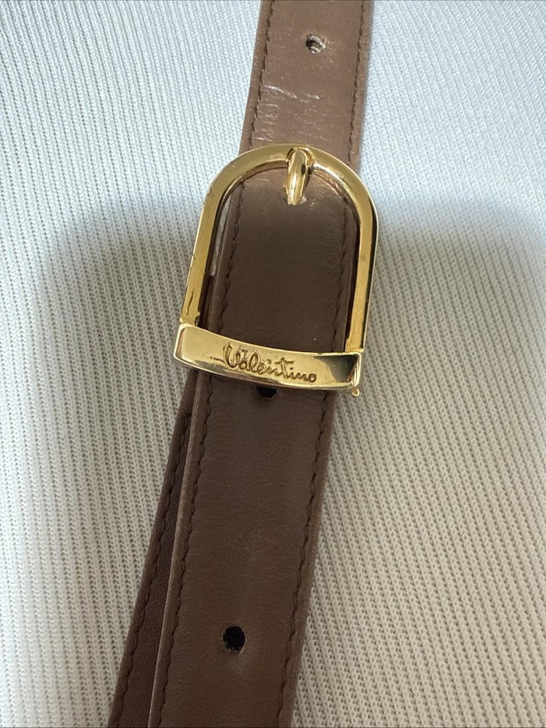 May include: A brown leather strap featuring a polished gold-tone metal buckle. The buckle is engraved with the word Valentino in a cursive script. The strap includes a single visible adjustment hole and a smooth, matte finish on the leather surface.