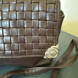 May include: A brown leather crossbody bag with a woven design. The bag features a long strap and decorative metal corner accents. The bag is made of a dark brown leather material.