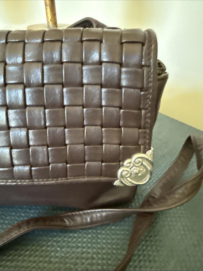 May include: A brown leather crossbody bag with a woven design. The bag features a long strap and decorative metal corner accents. The bag is made of a dark brown leather material.