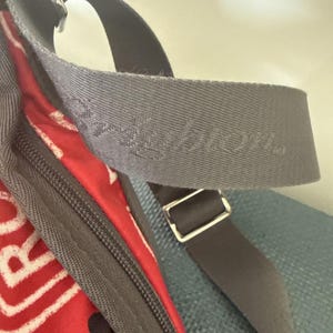 May include: Close-up of a gray shoulder strap with the embossed word "Brighton." The strap is part of a bag with a red and white patterned fabric visible. A dark gray strap with a silver buckle is also present.