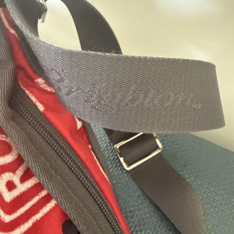 May include: Close-up of a gray shoulder strap with the embossed word "Brighton." The strap is part of a bag with a red and white patterned fabric visible. A dark gray strap with a silver buckle is also present.