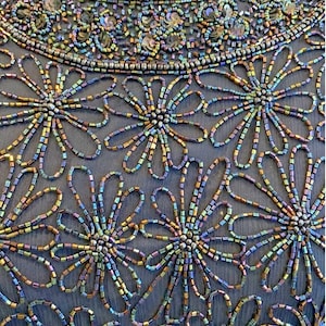 Op de afbeelding: Close-up van een formele jurk met ingewikkeld kralenwerk. De jurk heeft een donkerblauwe stoffen basis, versierd met iriserende kralen en pailletten. Bloempatronen worden gemaakt met de kralen en de halslijn is zwaar versierd.
