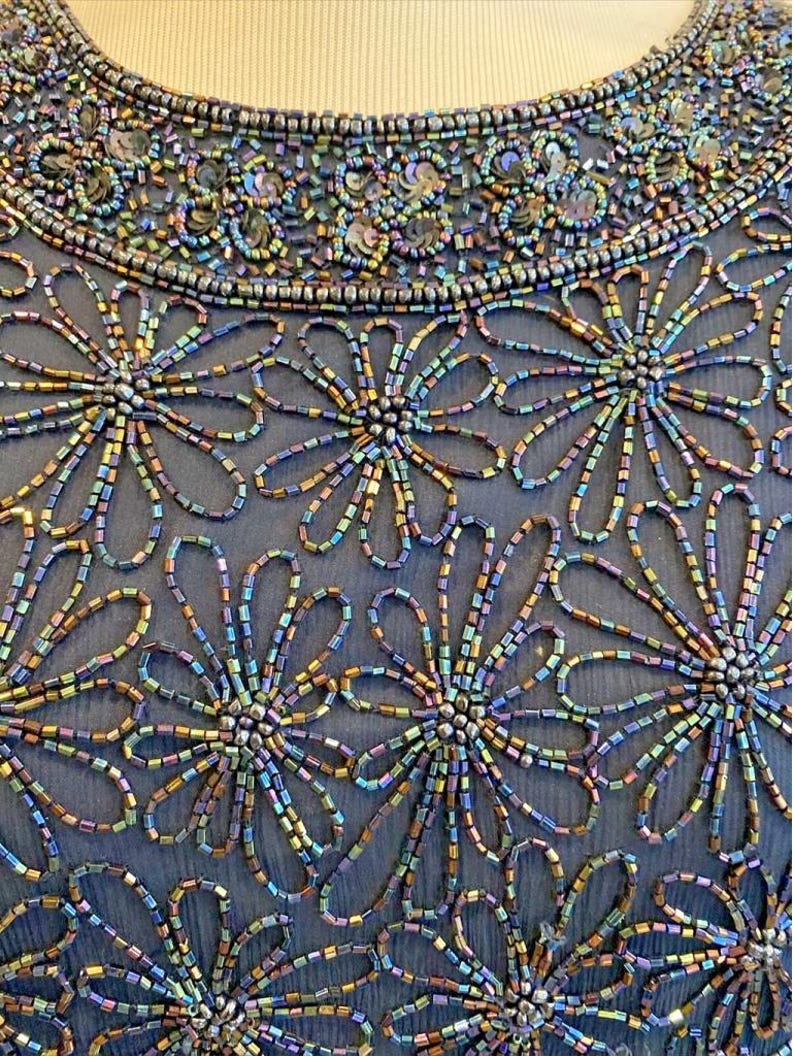 Op de afbeelding: Close-up van een formele jurk met ingewikkeld kralenwerk. De jurk heeft een donkerblauwe stoffen basis, versierd met iriserende kralen en pailletten. Bloempatronen worden gemaakt met de kralen en de halslijn is zwaar versierd.