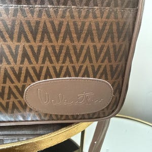 May include: A brown shoulder bag featuring a repeating V-shaped geometric pattern in a darker brown tone. The bag has a smooth, light brown oval-shaped leather patch on the front with the word Valentino embossed in a cursive script font.