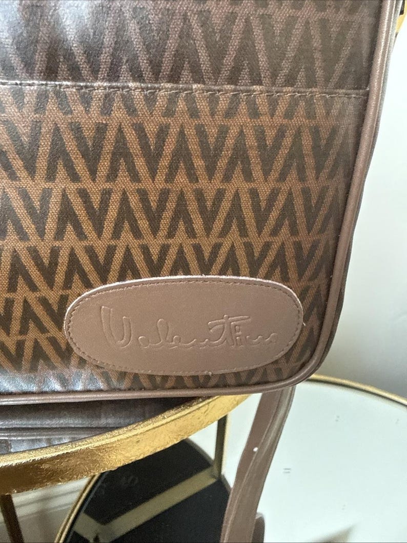 May include: A brown shoulder bag featuring a repeating V-shaped geometric pattern in a darker brown tone. The bag has a smooth, light brown oval-shaped leather patch on the front with the word Valentino embossed in a cursive script font.