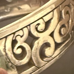 May include: A silver-toned metal bangle bracelet featuring a continuous openwork design of raised, swirling scroll patterns. The metallic surface has a matte finish with darker recessed areas that highlight the intricate, repeating curved motifs.