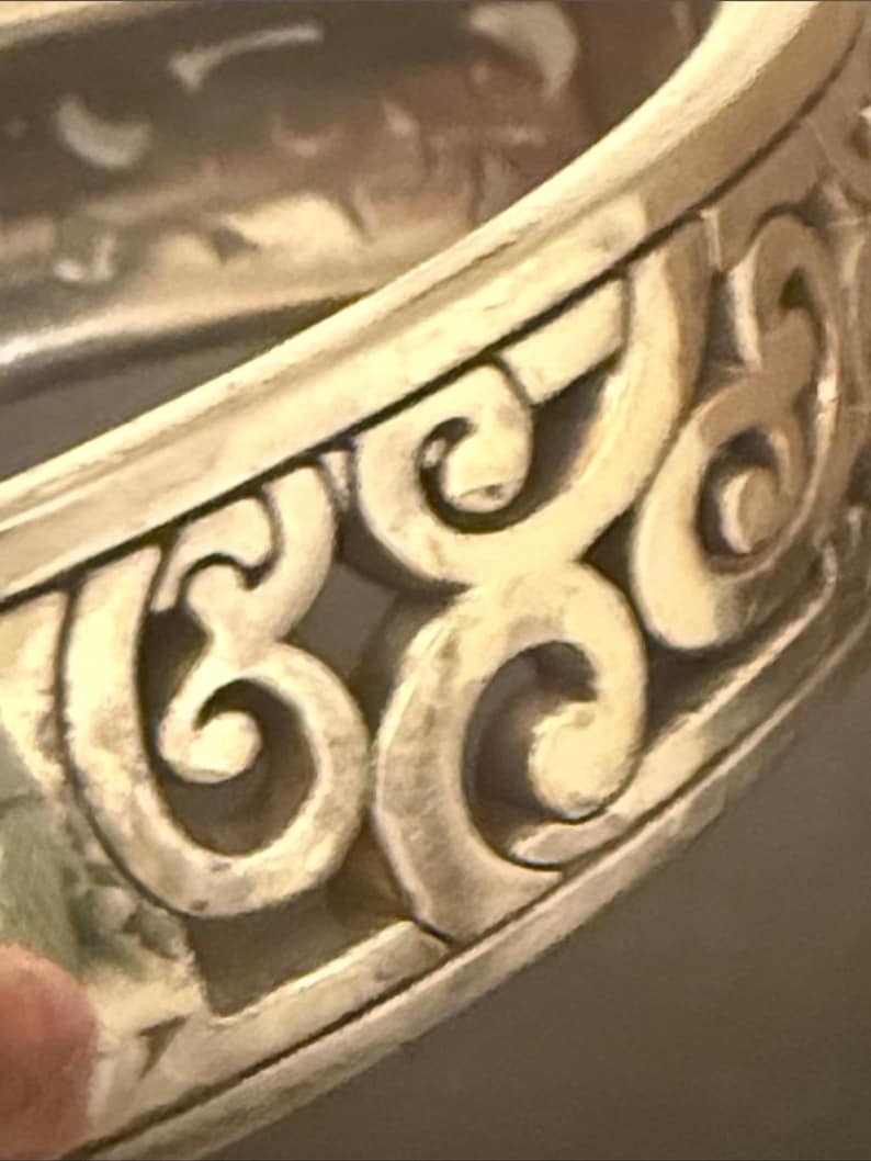 May include: A silver-toned metal bangle bracelet featuring a continuous openwork design of raised, swirling scroll patterns. The metallic surface has a matte finish with darker recessed areas that highlight the intricate, repeating curved motifs.
