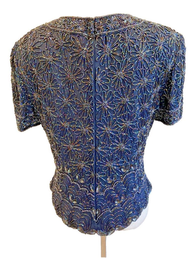 Op de afbeelding: Een marineblauwe, met kralen bezette top met korte mouwen. De top is bedekt met een bloemmotief van iriserende kralen. De halslijn en zoom zijn ook versierd met kralen. De achterkant van de top is zichtbaar, met een ritssluiting.