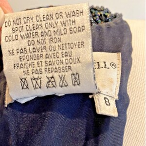 Op de afbeelding: Close-up van een marineblauw kledingstuk met een kralenrand. Een wit label toont wasvoorschriften in het Engels en Frans, waaronder "DO NOT DRY CLEAN OR WASH". Een ander label toont het nummer "8".