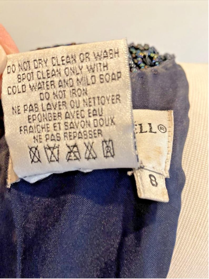 Op de afbeelding: Close-up van een marineblauw kledingstuk met een kralenrand. Een wit label toont wasvoorschriften in het Engels en Frans, waaronder "DO NOT DRY CLEAN OR WASH". Een ander label toont het nummer "8".