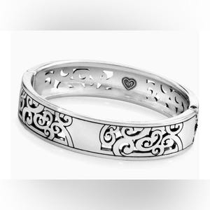May include: A silver-toned hinged bangle bracelet featuring an intricate cut-out scrollwork pattern along the outer band. The interior surface displays a small heart-shaped emblem with the letters JB engraved in the centre of the metal band.