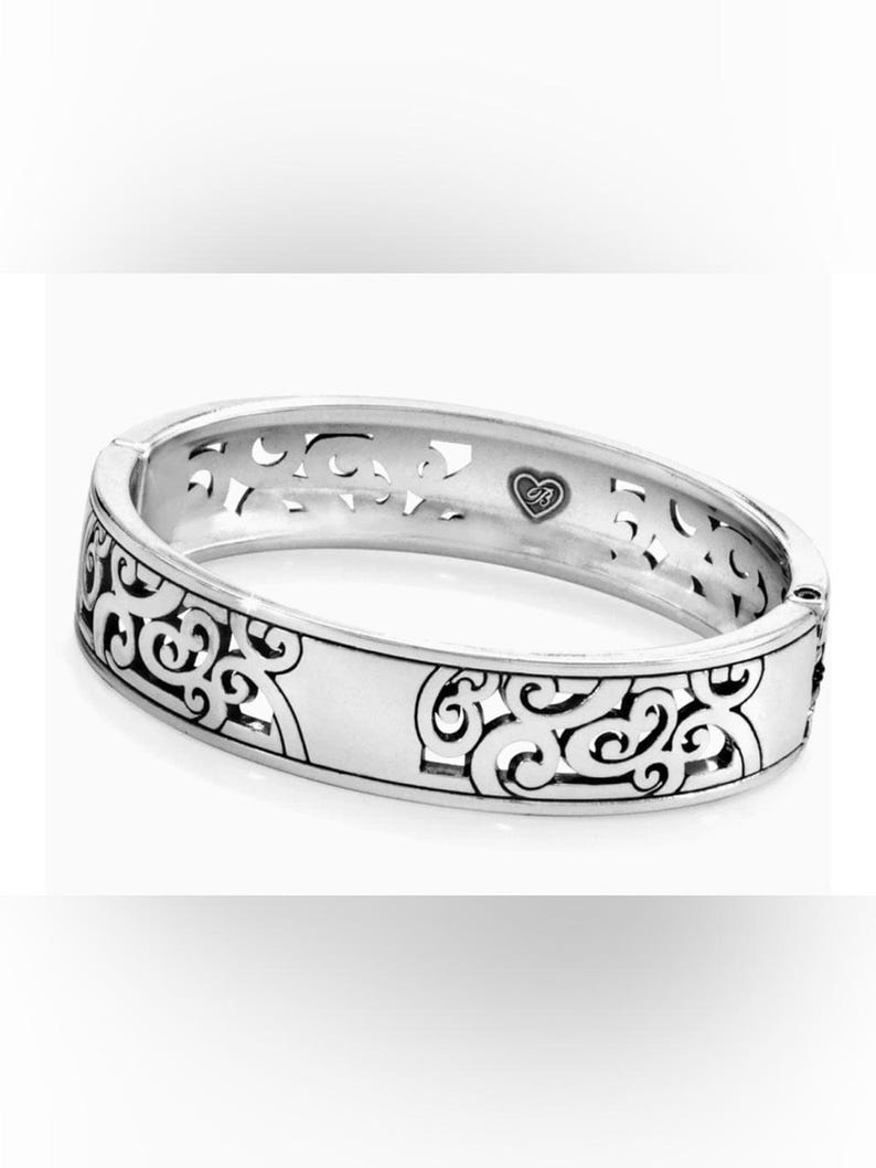 May include: A silver-toned hinged bangle bracelet featuring an intricate cut-out scrollwork pattern along the outer band. The interior surface displays a small heart-shaped emblem with the letters JB engraved in the centre of the metal band.