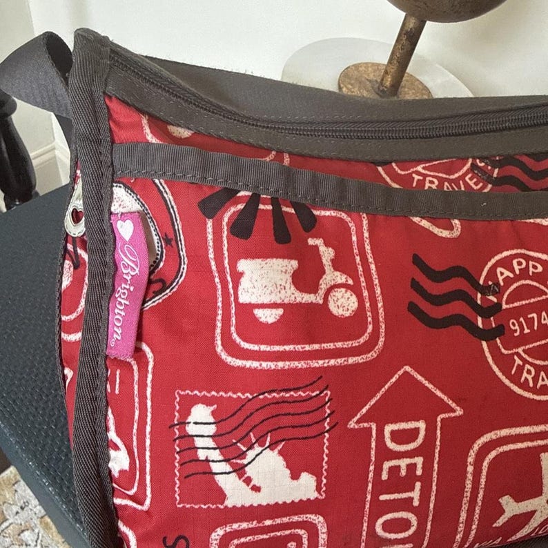 May include: Red crossbody bag with a travel-themed print featuring white scooter and stamp illustrations. The bag has a gray strap and trim, and a pink Brighton tag. The bag is designed for carrying essentials.