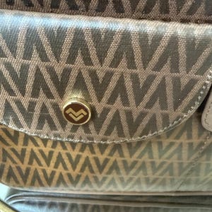May include: A brown handbag featuring a repeating geometric V-shaped pattern across the fabric. The front flap includes a circular gold-toned metal button closure with a stylized V logo in the center, providing a secure fastening mechanism.