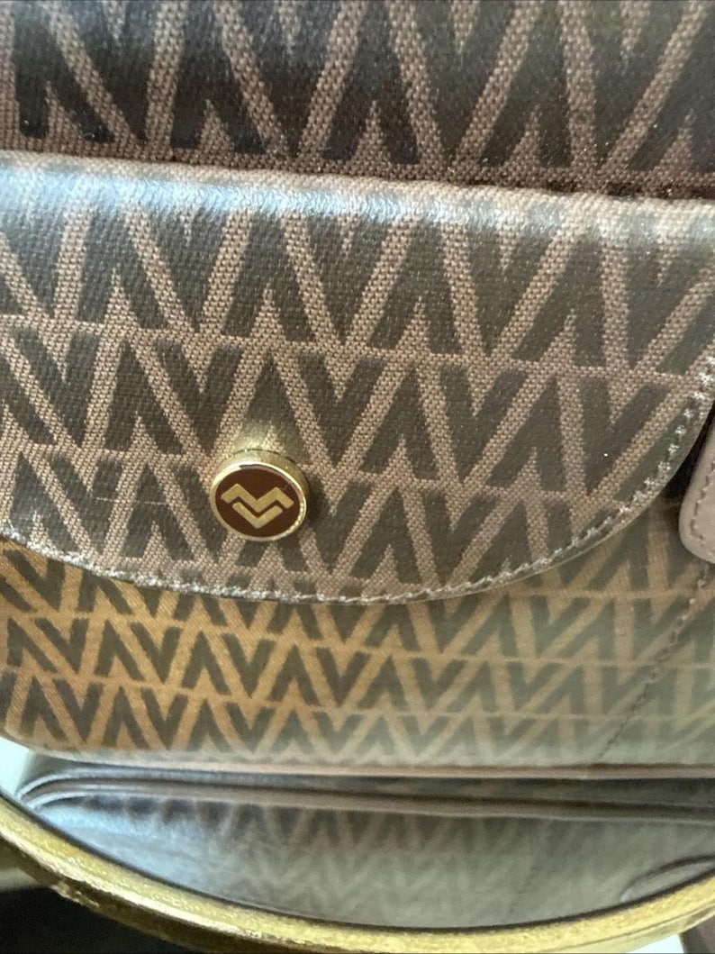May include: A brown handbag featuring a repeating geometric V-shaped pattern across the fabric. The front flap includes a circular gold-toned metal button closure with a stylized V logo in the center, providing a secure fastening mechanism.