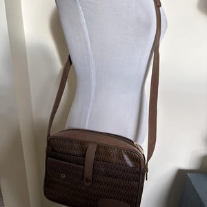 May include: A brown rectangular crossbody bag featuring a repeating geometric pattern across the front panel. The bag includes a central vertical strap with a snap closure, a front pocket, a top zipper, and an adjustable shoulder strap.