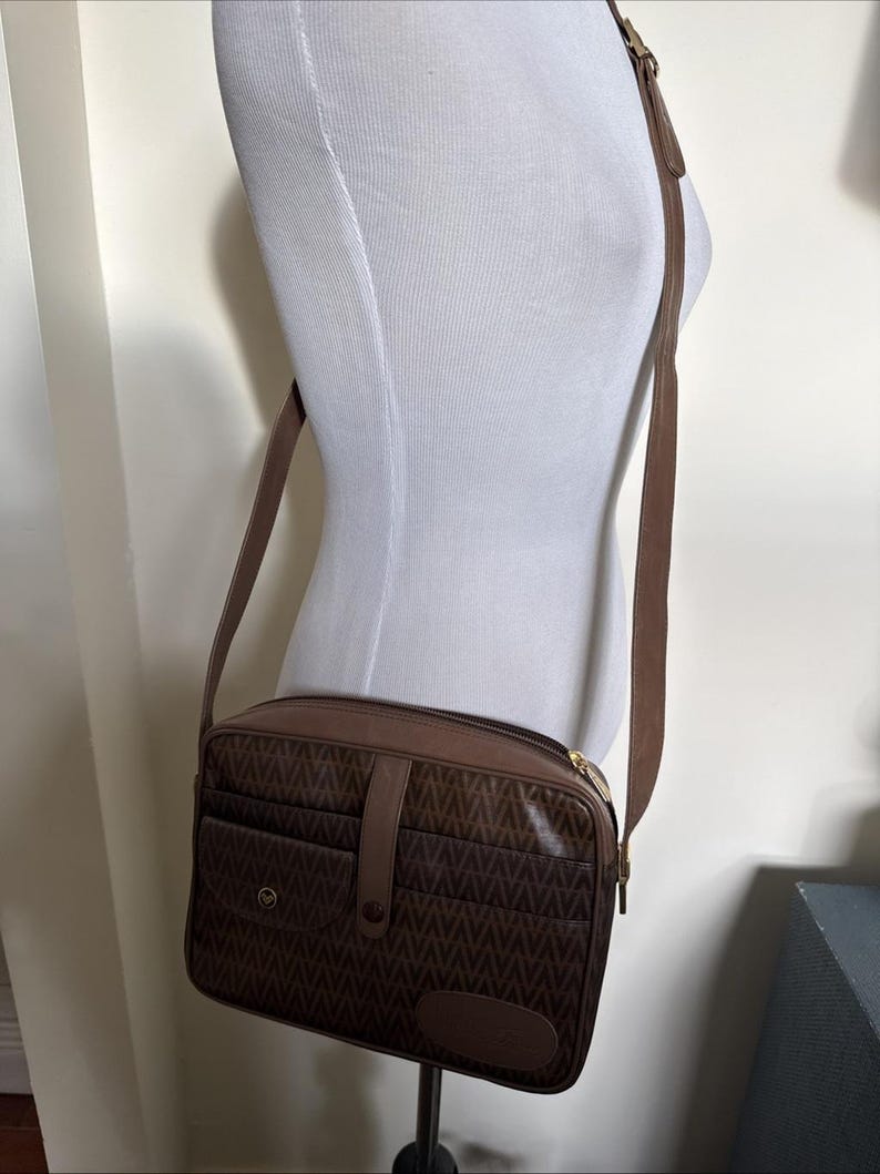 May include: A brown rectangular crossbody bag featuring a repeating geometric pattern across the front panel. The bag includes a central vertical strap with a snap closure, a front pocket, a top zipper, and an adjustable shoulder strap.