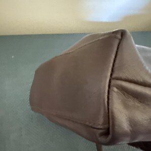 May include: A close-up of a taupe-colored leather handbag. The bag has a structured design with visible stitching and a strap. The leather appears smooth and has a slight sheen. The bag's corner is visible, showing its construction.