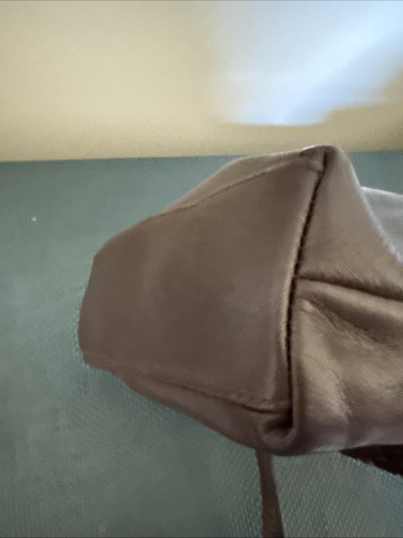 May include: A close-up of a taupe-colored leather handbag. The bag has a structured design with visible stitching and a strap. The leather appears smooth and has a slight sheen. The bag's corner is visible, showing its construction.