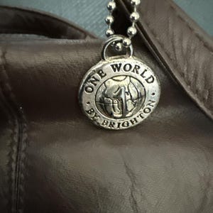 May include: Close-up of a brown leather handbag with a silver charm. The charm is round and features the words "ONE WORLD BY BRIGHTON" around a globe design. The charm hangs from a silver ball chain.
