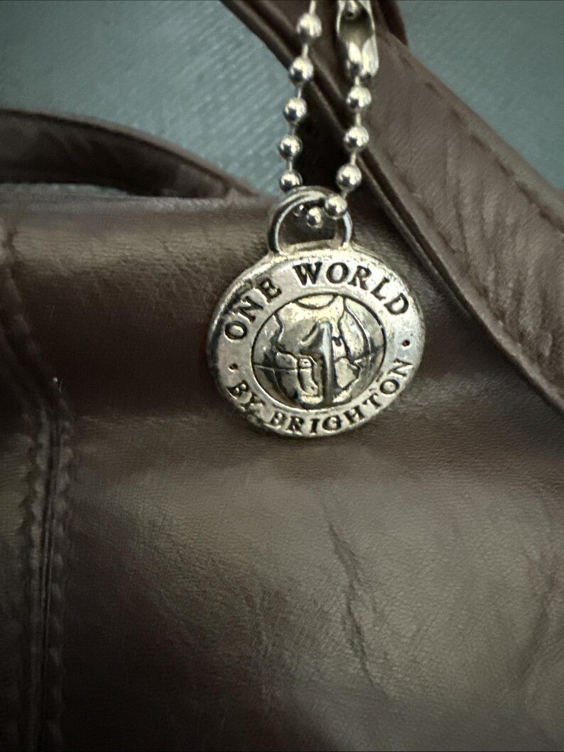 May include: Close-up of a brown leather handbag with a silver charm. The charm is round and features the words "ONE WORLD BY BRIGHTON" around a globe design. The charm hangs from a silver ball chain.