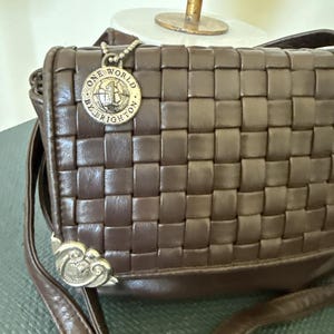 May include: A brown leather crossbody bag with a woven design. The bag features a silver-toned charm with the words "ONE WORLD BY BRIGHTON". The strap is brown and the bag has a silver-toned decorative clasp.