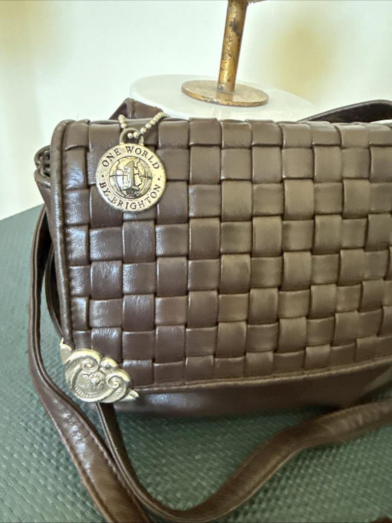 May include: A brown leather crossbody bag with a woven design. The bag features a silver-toned charm with the words "ONE WORLD BY BRIGHTON". The strap is brown and the bag has a silver-toned decorative clasp.