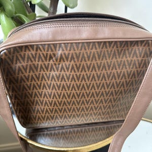 May include: A rectangular taupe leather shoulder bag featuring a central panel with a repeating dark brown V-shaped geometric pattern. The bag has a top zipper closure, smooth leather trim, and a matching leather shoulder strap attached to the sides.