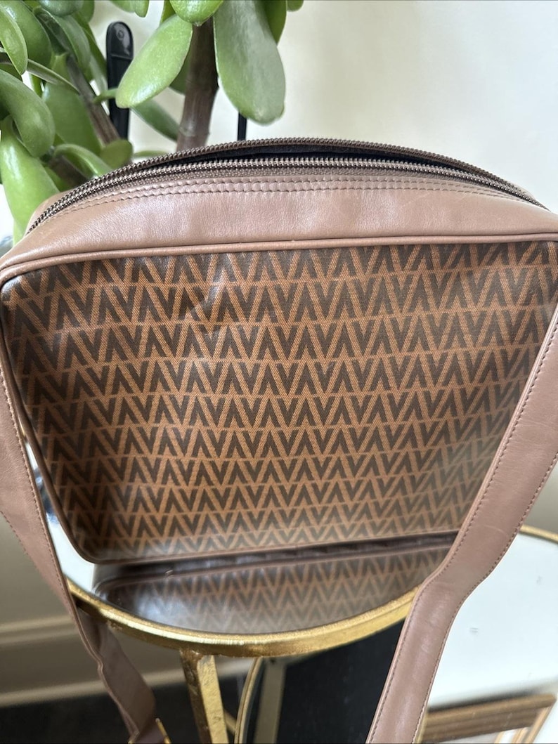 May include: A rectangular taupe leather shoulder bag featuring a central panel with a repeating dark brown V-shaped geometric pattern. The bag has a top zipper closure, smooth leather trim, and a matching leather shoulder strap attached to the sides.