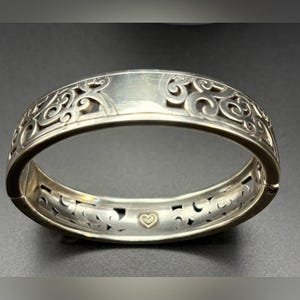 May include: A silver-toned hinged bangle bracelet featuring an intricate openwork scroll pattern along the outer band. The interior surface displays a smooth finish with a small, centred gold-toned heart emblem near the hinge mechanism.