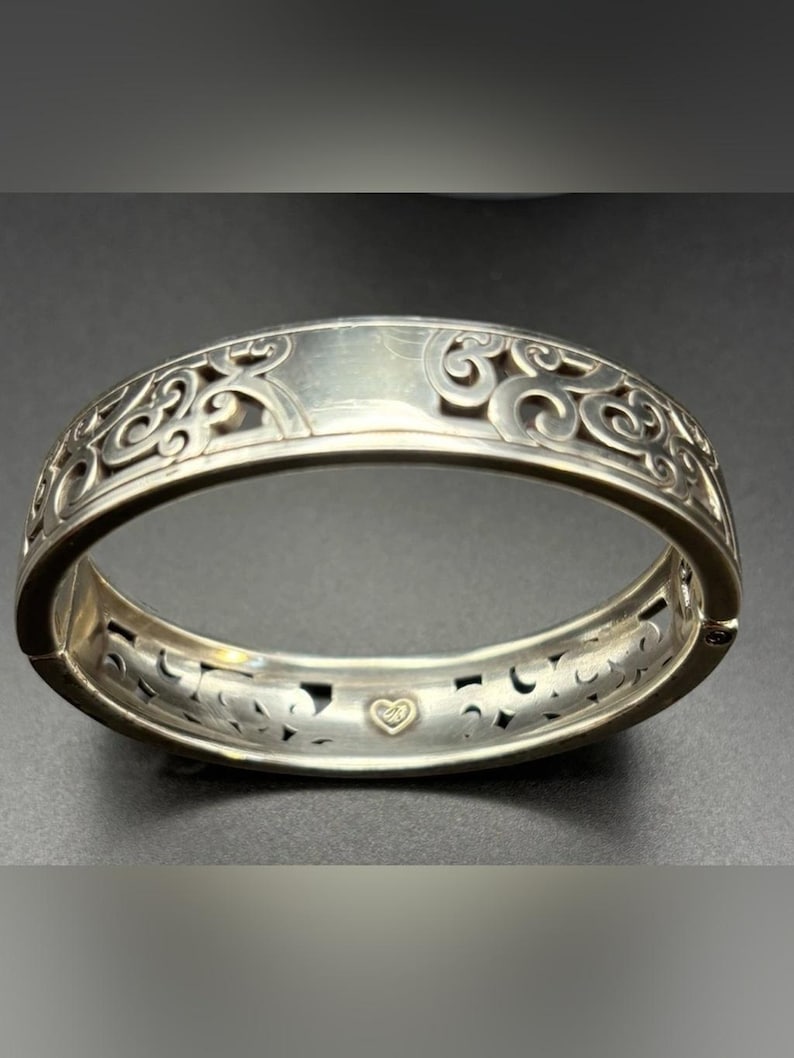 May include: A silver-toned hinged bangle bracelet featuring an intricate openwork scroll pattern along the outer band. The interior surface displays a smooth finish with a small, centred gold-toned heart emblem near the hinge mechanism.