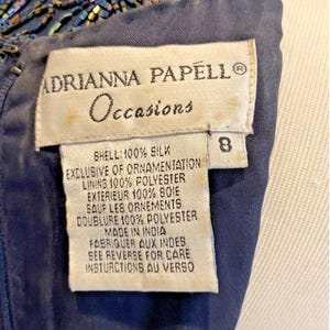 Op de afbeelding: Detailopname van een marineblauwe jurk met kralenversiering. Het label luidt "ADRIANNA PAPELL Occasions" en bevat de stofsamenstelling: 100% zijde voor de buitenkant, 100% polyester voor de voering. Maat 8.