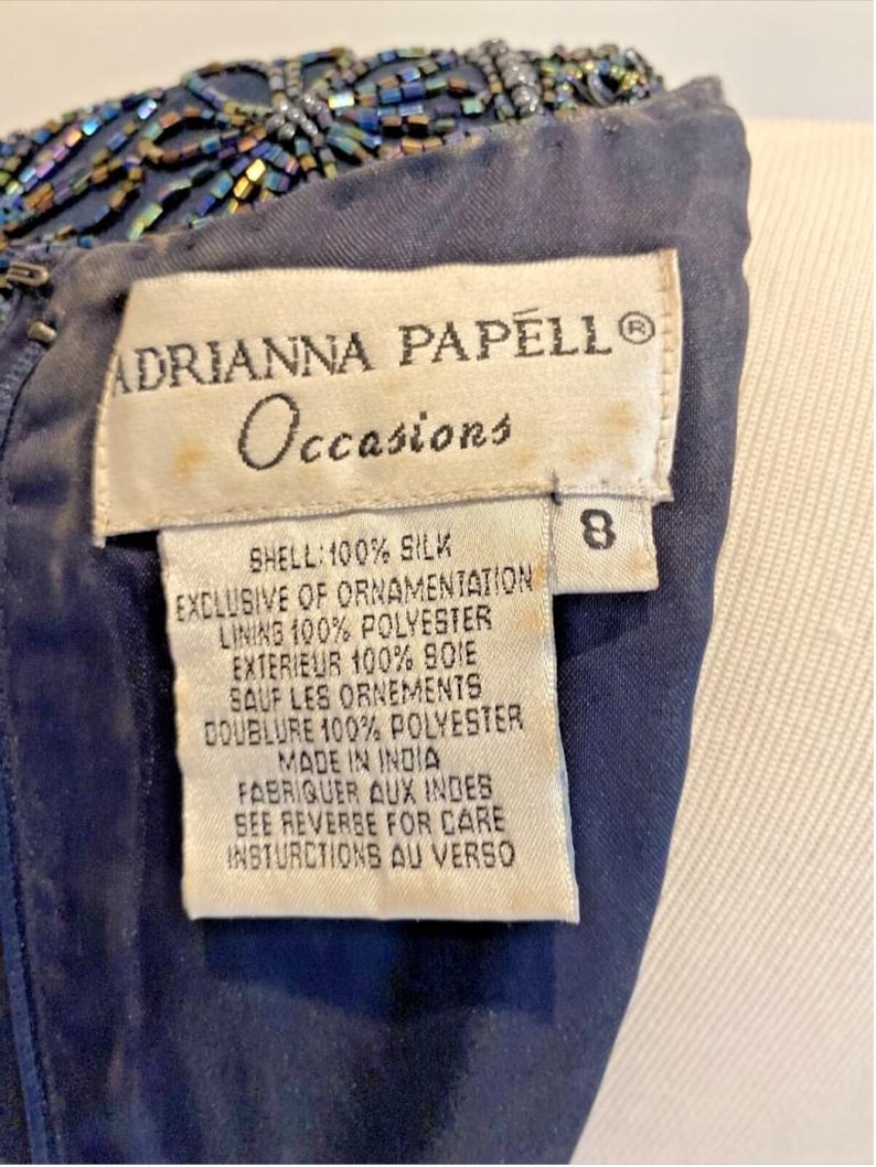 Op de afbeelding: Detailopname van een marineblauwe jurk met kralenversiering. Het label luidt "ADRIANNA PAPELL Occasions" en bevat de stofsamenstelling: 100% zijde voor de buitenkant, 100% polyester voor de voering. Maat 8.