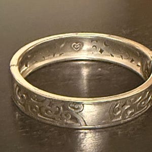 May include: A silver-toned hinged bangle bracelet featuring an embossed swirling pattern on the exterior surface. The interior of the metal band displays engraved celestial motifs including a small heart, crescent moons, and four-pointed stars.